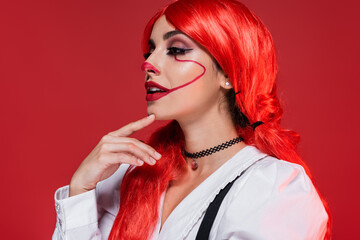 thoughtful and smiling woman with bright hair and clown makeup looking away isolated on red.