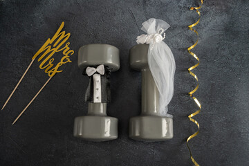 Two heavy gym dumbbells stylized as bride and groom, dressed in a small decorative veil and a suit or tuxedo. Healthy fitness lifestyle wedding ceremony concept, flat lay composition.	