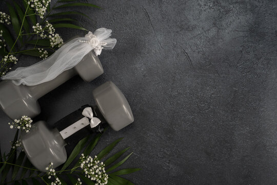 Two heavy gym dumbbells stylized as bride and groom, dressed in a small decorative veil and a suit or tuxedo. Healthy fitness lifestyle wedding ceremony concept, flat lay composition with copy space.