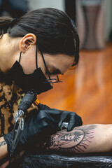 Detail of young tattoo artist girl with glasses and mask making a tattoo of 'La Santa Muerte' (Our Lady of Holy Death) in the arm of a woman with tattoo machine