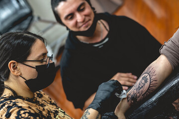Fototapeta na wymiar Detail of young tattoo artist girl with glasses and mask cleaning a new tattoo of 'La Santa Muerte' (Our Lady of Holy Death) in the arm of a woman and apprentice watching