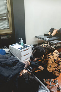 Young Tattoo Artist Girl With Glasses And Mask Making A Tattoo Of 'La Santa Muerte' (Our Lady Of Holy Death) In The Arm Of A Woman With Tattoo Machine