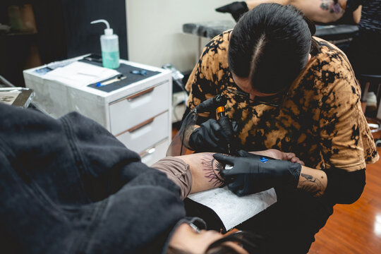 Young Tattoo Artist Girl With Glasses And Mask Making A Tattoo Of 'La Santa Muerte' (Our Lady Of Holy Death) In The Arm Of A Woman With Tattoo Machine