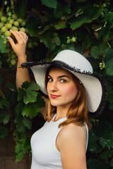 A woman in a vineyard holds grapes in her hand and smiles .in a white shirt and hat