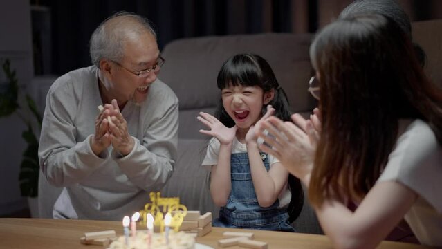 Asian happy family of little girl blowing out candles on cake. Celebrate birthday anniversary party with  Grandparents and mother on table at night in living room. Kid girl having happiness lifestyle.
