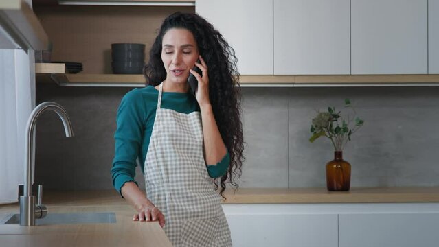 Pretty arabian curly haired woman mother housewife wearing apron stand in kitchen talk mobile phone friendly conversation calling to delivery food service make order from home spend free time alone