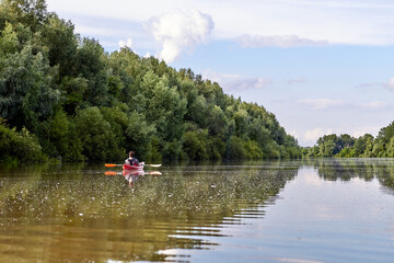 Man in red kayak on river near the shore with green trees against the blue sky and clouds