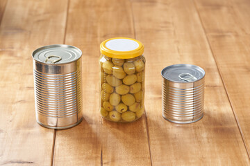 Iron tin can with tab opener and olives in a glass jar on the wooden table.