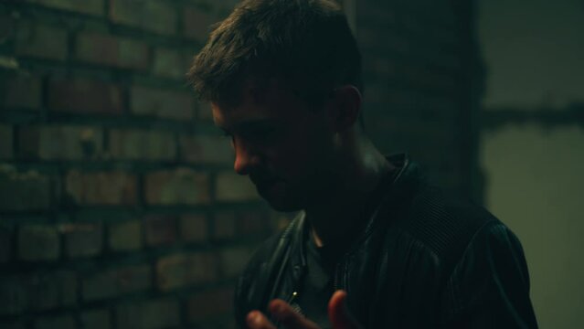 Mafia member or criminal in leather jacket after the murder touching his head with wound and leaving the crime scene. Flickering light in dark basement. Movie scene
