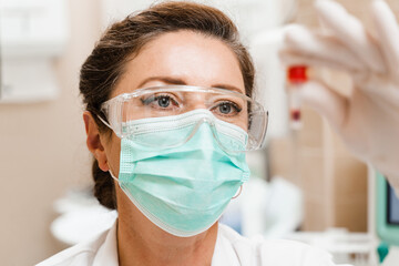 Laboratory assistant holds test tube with blood in her hands and looks at it. Blood test in the laboratory. Medical scientist in goggles and mask.