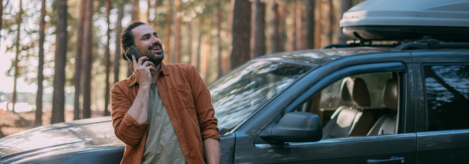 Obraz na płótnie Canvas A happy, joyful man is talking on the phone in the woods near the car on a sunny day.