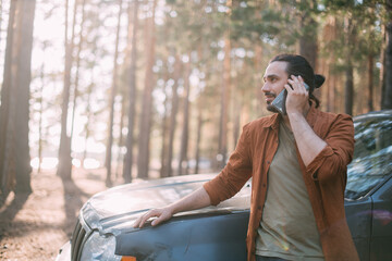 A happy, joyful man is talking on the phone in the woods near the car on a sunny day.