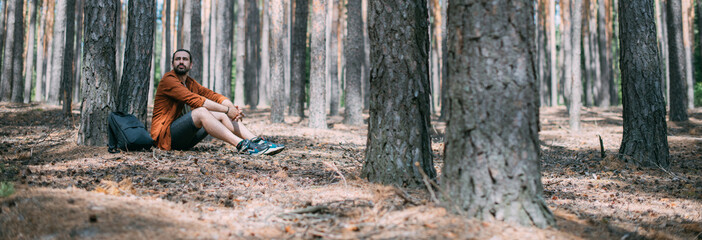 Portrait of a young man sitting at the foot of a pine tree in a coniferous forest on a sunny day. A...
