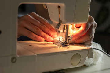 Hands of experienced worker in the handmade industry, sewing white fabric on a sewing machine, the production of Santa red hat. Homemade sewing. Working tailor. Christmas hat. Close up.