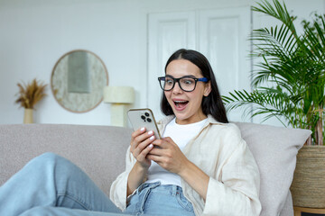 Happy young woman in glasses holding mobile phone, celebrating unexpected victory in game, won....