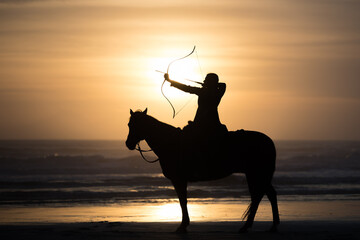 Mounted archer holds bow and arrow at sunrise on the beach. - obrazy, fototapety, plakaty