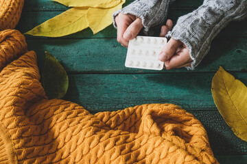 Female hands holding a blister of pills, autumn flu, epidemic concept, yellow fall leaves and warm knitted sweater on backdrop
