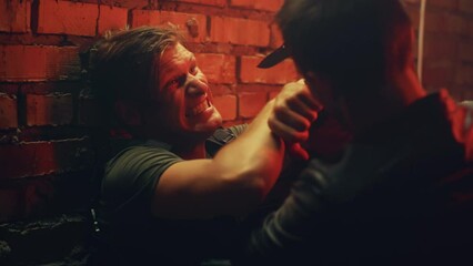 Male bandit or mafia member pressing police officer into the brick wall and attacking him with knife on dim street at night. Action or criminal movie cinematic scene with creative lighting