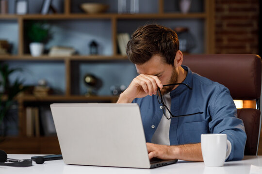 Handsome male businessman manager working tiredly behind a laptop in a stylish home office. Loss of vision from the screen. Fatigue and burnout from work. Overwork and stress.