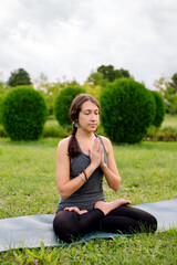 Naklejka na ściany i meble Front view of a woman instructor sitting on yoga mat in lotus position outside in city park. Namaste mudra, seated yoga asana, lotus pose, lotus posture, padmasana, cross legged sitting posture.