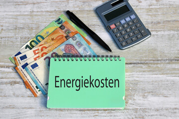 Grüner Notizblock mit dem Wort Energiekosten mit Eurobanknoten, Taschenrechner und Kugelschreiber.
