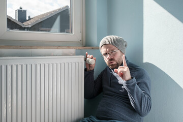 Man in warm clothes sits near the radiator and turns the thermostat to the minimum, making a...