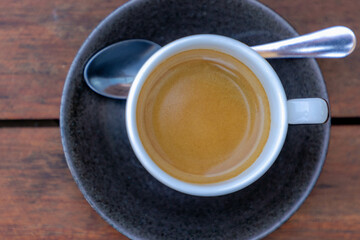Selective focus on white coffee cup and saucer and spoon on the wooden table, Top view of black coffee and brown creamy foam with free copy space, Outdoor drinking warm drink in the afternoon.