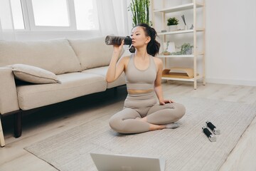 Sporty Asian woman in a yoga suit blogger watching a recording of a sports workout on her laptop smiling and drinking water while relaxing at home, lifestyle in sports and good nutrition