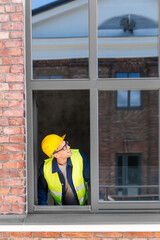 construction business and building concept - smiling male builder in helmet and safety west looking out window