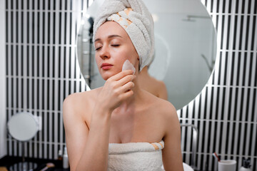Young beautiful woman wearing white towel doing face massage with pink quartz scraper during beauty morning routine in bathroom. Anti aging treatments and skincare concept