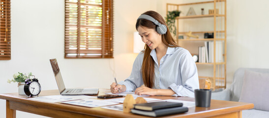 Female freelance is reading business data on laptop and writing information on notebook while wearing