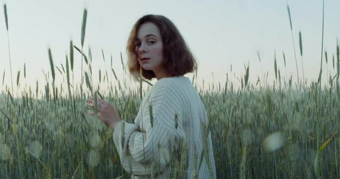 A young red-haired woman turns her head and looks directly into the camera while standing among tall ears in a wheat field. 4k video, red komodo