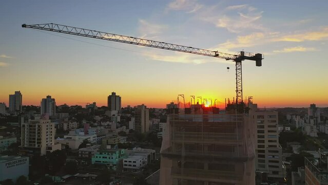 Drone sobrevoando cidade ao p&ocirc;r do sol, foco em um pr&eacute;dio sendo constru&iacute;do com uso de grua. Cidade ao fundo ao entardecer.