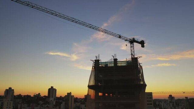 Drone sobrevoando cidade ao p&ocirc;r do sol, foco em um pr&eacute;dio sendo constru&iacute;do com uso de grua. Cidade ao fundo ao entardecer.