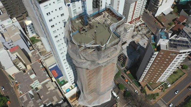 Drone sobrevoando um pr&eacute;dio sendo constru&iacute;do com uso de grua e homens ao longe trabalhando. Lage sendo executada. Cidade ao fundo.