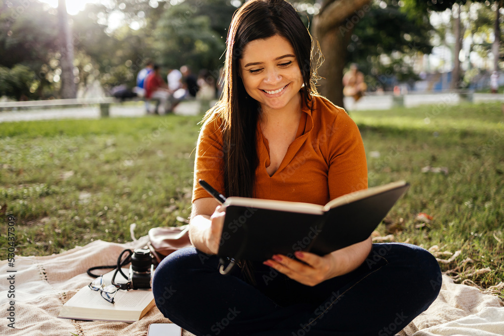 Wall mural Portrait of young woman sitting on green grass in park during summer day and writing notes in notebook with pen