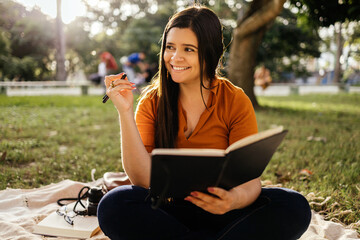 Portrait of young woman sitting on green grass in park during summer day and writing notes in...