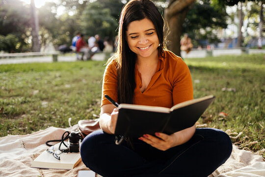 Portrait of young woman sitting on green grass in park during summer day and writing notes in notebook with pen