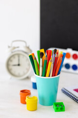 Obraz premium Time to return back to school. Bright colourful green yellow, blue stationery, paints, pencils, markers, brushes. black board on background. White alarm clock. Copy space for text. Autumn, September