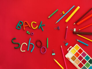words of multi-colored plasticine back to school in wry funny style and school supplies felt-tip pens, paints, pen, paper clips, on red background top view