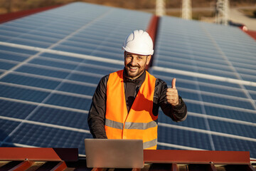 A happy supervisor is checking on solar panels on the laptop and giving thumbs up while smiling at the camera.