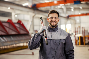 A heavy industry worker poses with big industrial pliers on his shoulder and smiles at the camera.