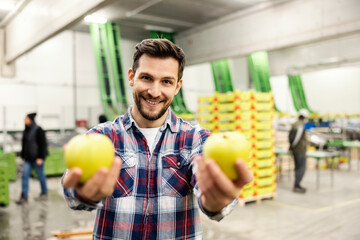 A happy fruit factory worker offering apples while standing in storage.