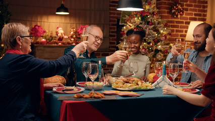 Happy senior man inviting family members to toast while celebrating winter holiday at home. Festive relatives gathered to celebrate Christmas with traditional home cooked food and sparkling wine.