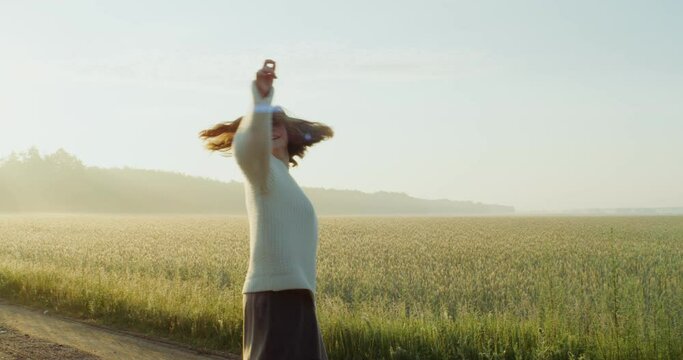A beautiful young red-haired woman laughs as she dances and whirls on a gravel road near an agricultural field, enjoying the warm rays of the rising sun. 4k, video, red komodo