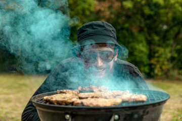 man in a hat smiles from behind thick smoke while grilling meat