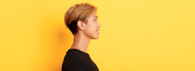 Profile of stylish handsome asian guy with fair hair looking left and smiling, standing over yellow...