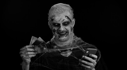 Sinister man with horrible scary Halloween zombie make-up using credit bank cards and smartphone while transferring money purchases online shopping. Dead guy with wounded bloody scars face, black room