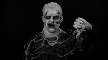 Sinister man in carnival costume of Halloween crazy zombie with bloody wounded scars face showing thumbs down sign gesture, expressing discontent, disapproval, dissatisfied, dislike. Cosplay undead