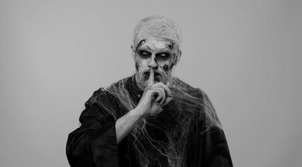 Shh be quiet. Sinister man with horrible scary Halloween zombie makeup in costume presses index finger to lips makes silence gesture sign do not tells secret. Dead guy with wounded bloody scars face - obrazy, fototapety, plakaty
